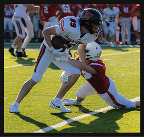 Two people in football uniforms are engaged in a tackle on a sunlit field with one person holding the ball and the other reaching to stop them. Description generated by AI