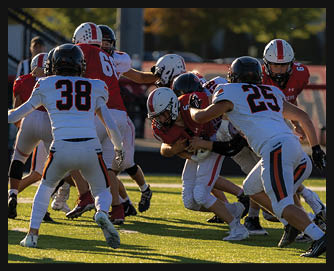 Several people wearing football uniforms are engaged in a tackle on a sunlit field during a game. Description generated by AI