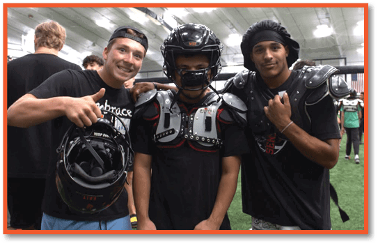Three people in a sports facility, one smiling and giving a thumbs up while holding a helmet, another wearing football shoulder pads and a helmet, and the third standing close by. Description generated by AI