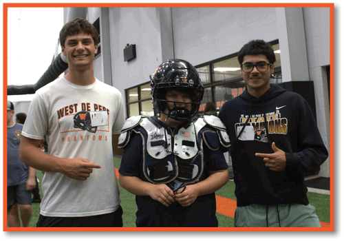 Three people stand indoors on artificial turf with one wearing football shoulder pads and a helmet while the others wear casual clothing and smile. Description generated by AI