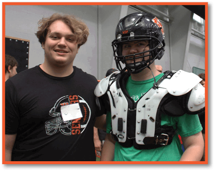 Two people are smiling, one wearing a black t-shirt with a football graphic and the other wearing football shoulder pads and a helmet indoors. Description generated by AI