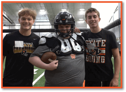 Three people stand together on an indoor sports field, with one person wearing football gear and holding a football while the others smile beside them. Description generated by AI