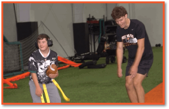 A person wearing headphones and a sports jersey is holding a football on an indoor turf field while another person stands nearby. Description generated by AI