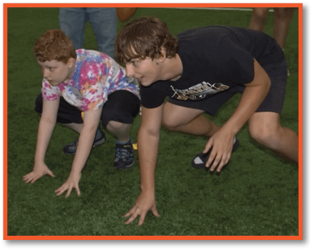 A person wearing a black t-shirt and shorts is crouched on a green indoor field, appearing ready to start a play, while another person nearby holds a football. Description generated by AI