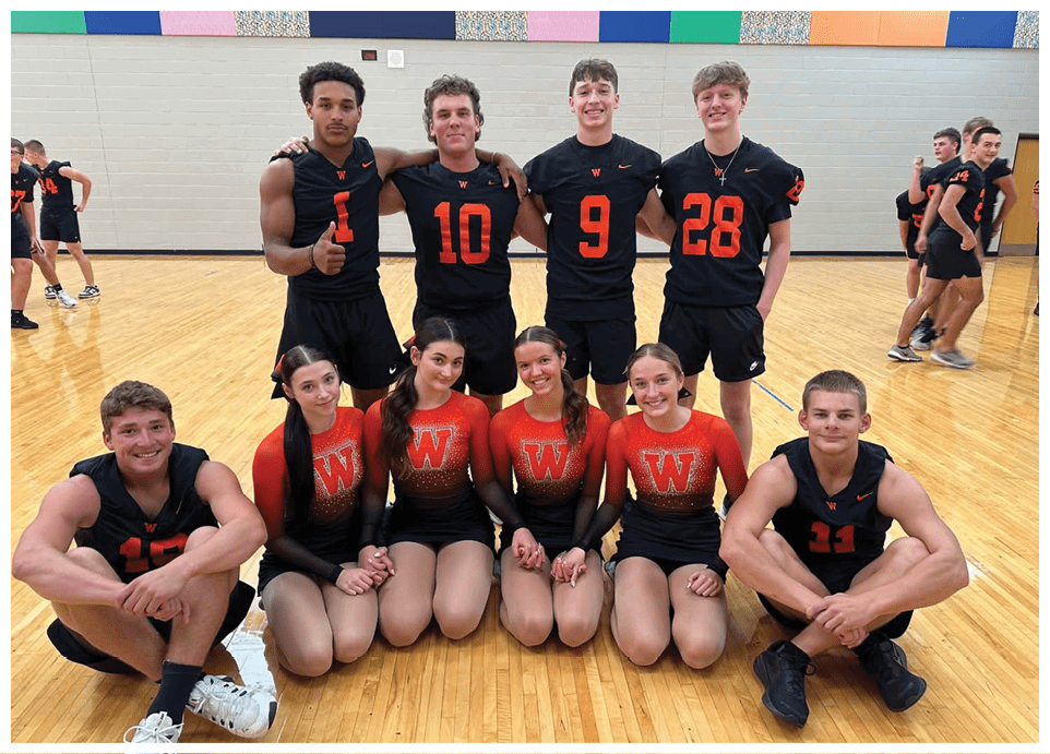 A group of people wearing matching sports uniforms with numbers and letters pose together on a gym floor, some standing with arms around each other and others kneeling in front holding hands. Description generated by AI