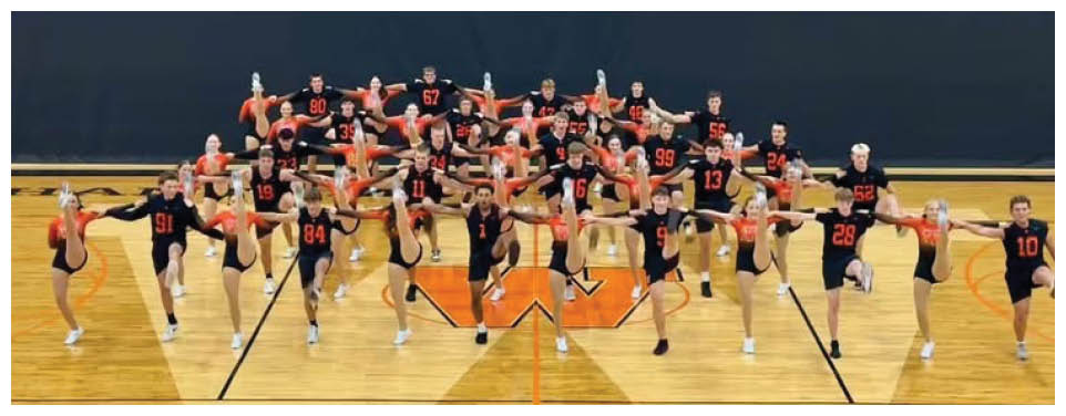 A group of people wearing matching sports uniforms with numbers and letters pose together on a gym floor, some standing with arms around each other and others kneeling in front holding hands. Description generated by AI