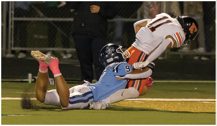 A person in a blue sports uniform tackles another person in a white and orange football uniform near the end zone on a football field while spectators watch behind a fence. Description generated by AI