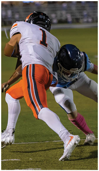 An adult wearing an orange and white football uniform with the number one is running on a field while being tackled by another adult in a white and blue uniform. Description generated by AI
