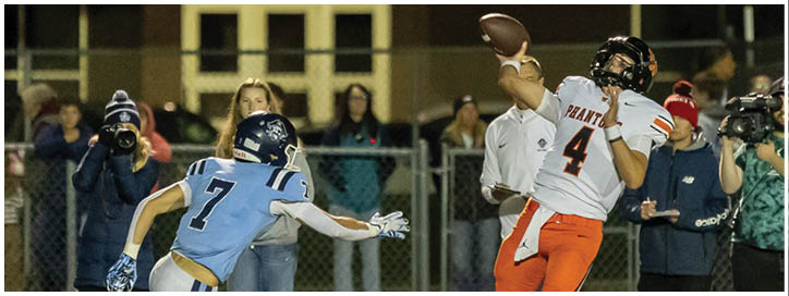 A person in a light blue sports jersey with the number seven is reaching out toward another person wearing orange sports pants on a field with a chain-link fence and spectators in the background. Description generated by AI