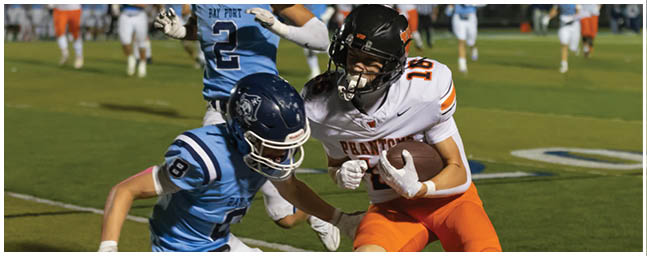 Two people in football uniforms are actively competing for the ball on a grassy field during a game at night. Description generated by AI