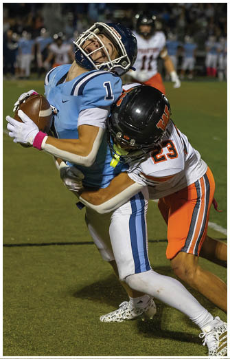 Two people in football uniforms are engaged in a tackle on a grassy field during a game, with one person wearing a blue and white uniform and the other in an orange and white uniform. Description generated by AI