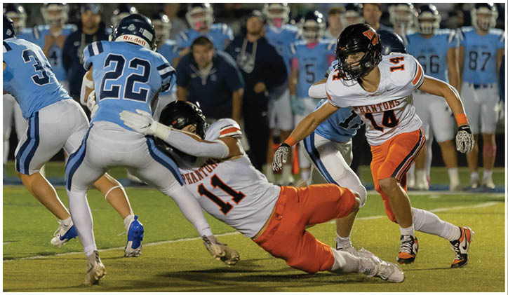 Two football players in white and orange uniforms tackle a player in a blue uniform during a game on a grassy field with other players and spectators in the background. Description generated by AI