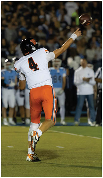 A person wearing a white and orange football uniform with the number 4 is throwing a football on a field with blurred spectators and players in the background. Description generated by AI