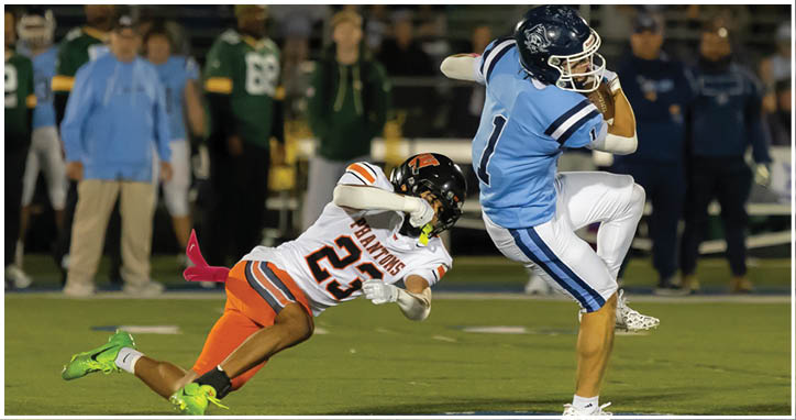 A person in a light blue football uniform is running while another person in an orange and white uniform is diving to tackle them on a football field. Description generated by AI