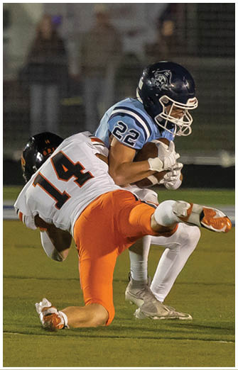 A person wearing a white and orange football uniform is tackling another person in a blue and white football uniform who is holding a football on a field. Description generated by AI