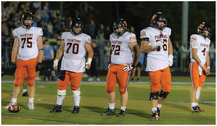 Five people wearing football uniforms with helmets stand on a grassy field, appearing to prepare for a play. Description generated by AI