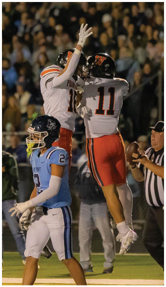 Two people in white and orange football uniforms jump to celebrate while a person in a blue and white uniform walks nearby on a football field with a crowd in the background. Description generated by AI