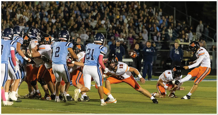 A group of people in football uniforms are on a field with one person kicking a ball held by another person while others prepare to block or tackle. Description generated by AI