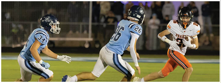 Two people in light blue football uniforms are running toward a person in an orange and white football uniform who is holding a football on a field. Description generated by AI