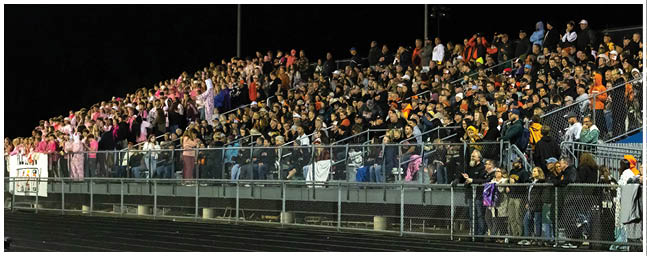 A large crowd of people wearing various colorful clothing is seated closely together on bleachers at an outdoor nighttime event. Description generated by AI