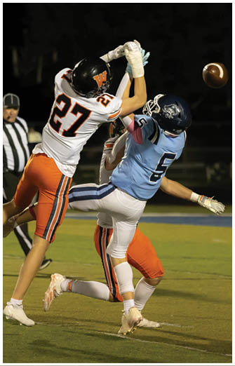 Two people in football uniforms are jumping and reaching for the ball during a nighttime game while a referee watches in the background. Description generated by AI