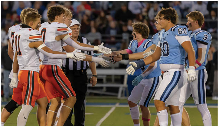 Players from two opposing football teams wearing different colored uniforms are shaking hands on the field with a referee standing nearby and a crowd in the background. Description generated by AI
