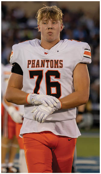 A person wearing a white football jersey with the number 76 and the word “PHANTOMS" stands on a field with hands clasped and wearing white gloves. Description generated by AI