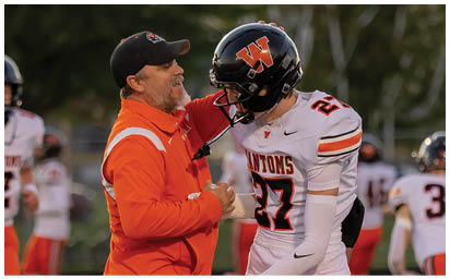 A person in an orange jacket and cap is smiling and interacting closely with a person in a white football uniform and black helmet on a sports field. Description generated by AI