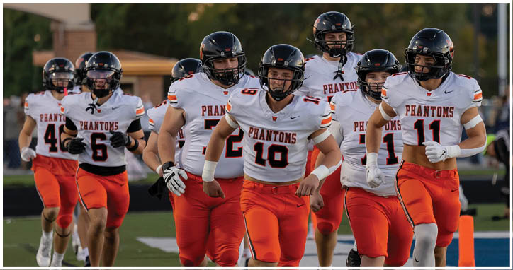 A group of adults wearing football uniforms with helmets and orange pants are running together on a field during a game or practice. Description generated by AI