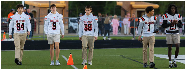 Five people wearing white sports jerseys with numbers walk together outdoors near a building with a peaked roof and trees in the background. Description generated by AI