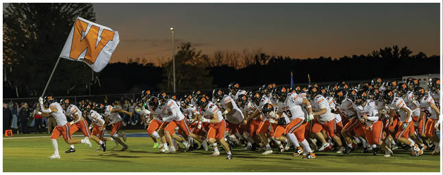 A group of people wearing helmets and uniforms are gathered outdoors at dusk with a large flag featuring a bold orange “W" waving above them. Description generated by AI
