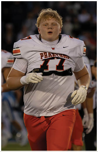A person wearing a white football jersey with the number 77 and red pants is walking on a field during a game. Description generated by AI