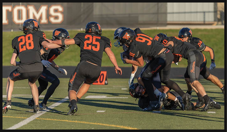 Several people wearing black and orange football uniforms are engaged in a play on a football field with one person on the ground being tackled by others. Description generated by AI