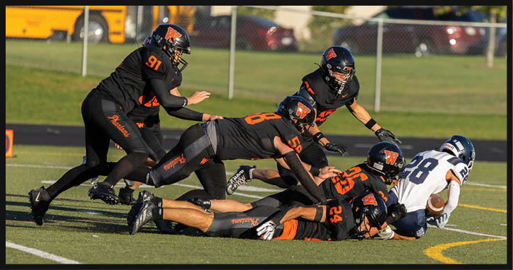 Several people in black football uniforms are tackling a person in a white football uniform who is holding a football on a grassy field. Description generated by AI