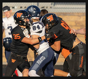 People wearing black and orange football uniforms are engaged in a play against opponents in white and blue uniforms on a grassy field. Description generated by AI
