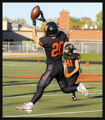 A person wearing a black and orange football uniform with the number 20 is jumping to catch a football on a green field while another person in a similar uniform stands in the background. Description generated by AI