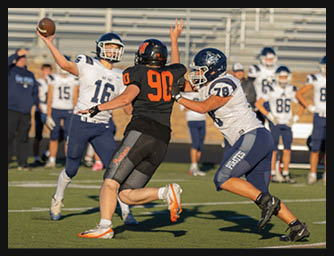 A person in a white and blue football uniform is throwing a football while another person in a black and orange uniform is running toward them and a third person in a white and blue uniform is attempting to block the runner on a football field. Description generated by AI