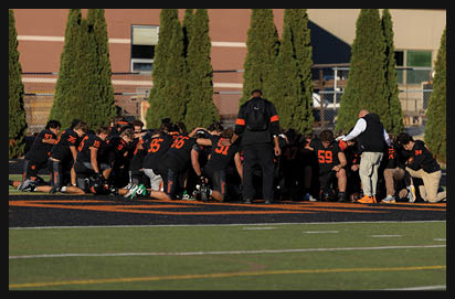 A group of people wearing black sports uniforms are kneeling on a field with one person standing and another person in light-colored pants and a vest also kneeling, appearing to be in a moment of reflection or prayer. Description generated by AI