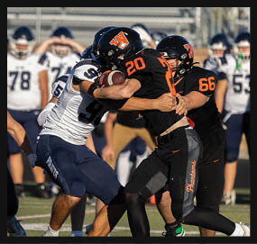 Two football players in black uniforms are tackling a player in a white uniform holding a football during a game on a grassy field. Description generated by AI