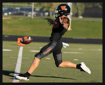 A person wearing a black football uniform with orange accents is kicking a football on a green field during daylight. Description generated by AI