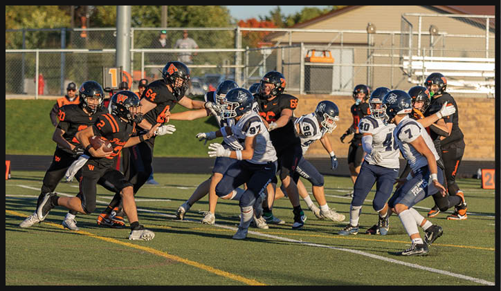 Adults in football uniforms are actively engaged in a game on a grassy field with a fence and trees in the background. Description generated by AI