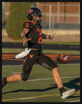 A person wearing a black football uniform with orange accents is kicking a football on a green field during daylight. Description generated by AI