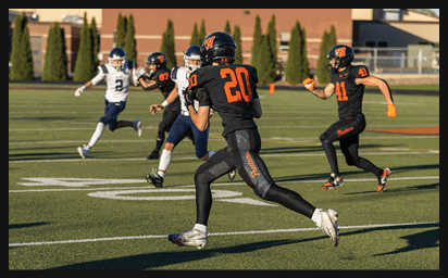 People in football uniforms are actively running on a field during a game, with some players in black and orange and others in white and blue. Description generated by AI