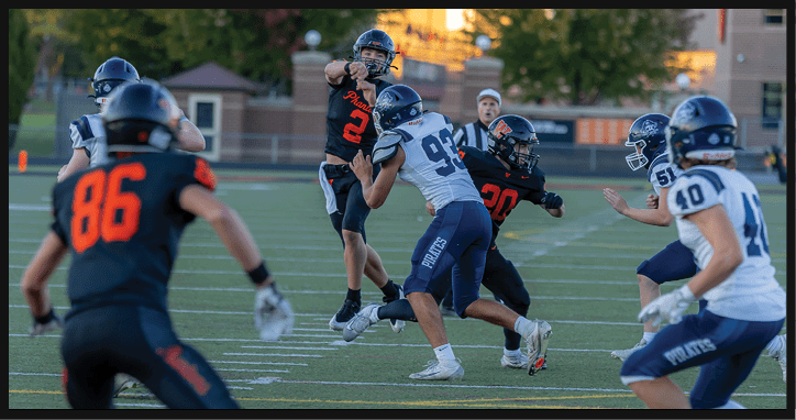 Several people wearing football helmets and uniforms are actively engaged in a play on a football field with trees and buildings in the background during daylight. Description generated by AI
