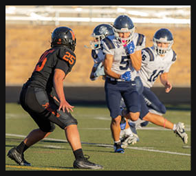 A person in a black football uniform with the number 25 faces three people in white and blue football uniforms during a game on a field. Description generated by AI