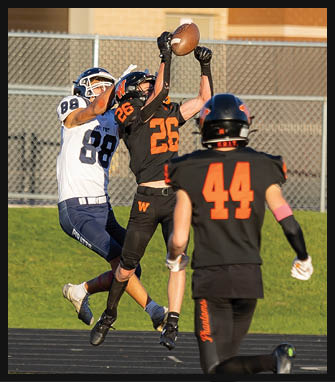 Two people in football uniforms are jumping and reaching to catch a football during a game on a field. Description generated by AI