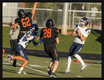 Two people in black football uniforms with orange numbers and one person in a white and blue football uniform are engaged in a play on a football field. Description generated by AI
