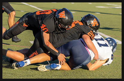 Three football players in black and orange uniforms tackle a player in a white and blue uniform on a grassy field with a school bus and an official in the background. Description generated by AI