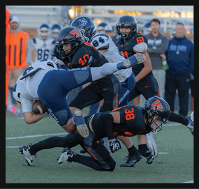 Several football players in black and white uniforms are engaged in a tackle on a green field with spectators in the background. Description generated by AI