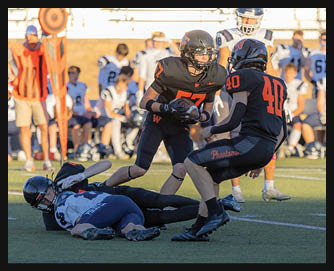 Two people in black football uniforms with orange numbers are engaged in a play on a football field while two people in white and blue uniforms are on the ground and others watch from the sidelines. Description generated by AI
